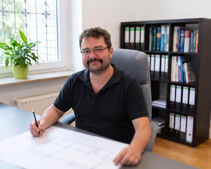 Gerd Johannes Bommes sitzt mit Bleistift über einem Bauplan an seinem Schreibtisch und schaut freundlich in die Kamera. Mit KI erstellt.